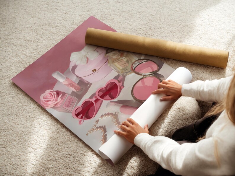 May include: A rolled-up print featuring a pink illustration of beauty items, including heart-shaped sunglasses, a perfume bottle, and a rose. A brown cardboard tube rests above the print. The print is on a white carpet.