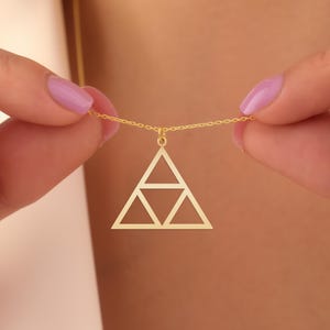May include: A gold-colored pendant necklace featuring a geometric triangle design. The pendant is suspended from a delicate chain. The necklace is held by a person with painted nails.