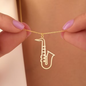 May include: A gold-colored necklace featuring a saxophone pendant. The pendant is a detailed cutout of the instrument, suspended from a delicate chain. The necklace is held by a person with painted nails.