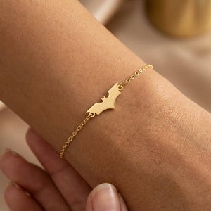 May include: A gold-colored bracelet featuring a Batman logo charm. The charm is a stylized bat silhouette, connected to a delicate chain. The bracelet is worn on a wrist, showcasing the jewelry's design and color.