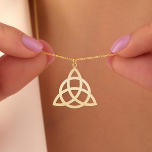May include: A gold-colored necklace featuring a Celtic knot pendant. The pendant is a stylized triangular shape with interwoven lines. The necklace is held by a person with painted nails. The chain is delicate and thin.