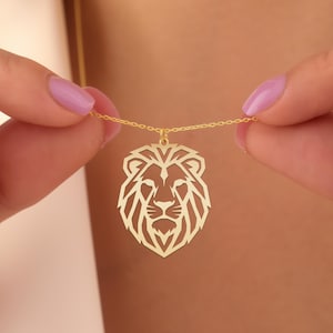 May include: A gold-colored necklace featuring a geometric lion pendant. The pendant is a detailed cutout of a lion's head, suspended from a delicate chain. The necklace is held between two hands with painted nails.