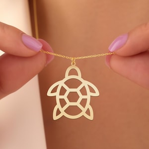 May include: A gold-colored turtle pendant necklace. The pendant is a cutout design with a turtle shape, including the head, flippers, and shell. The necklace chain is delicate and gold-toned. The necklace is being held up by a person.