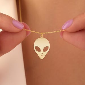 Könnte beinhalten: Eine goldfarbene Halskette mit einem Anhänger in Form eines Alienkopfes. Der Alien-Anhänger hat große ovale Augen und einen kleinen Mund. Die Halskette wird von einer Person mit rosa Nagellack gehalten.
