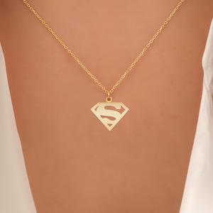 May include: A gold-colored necklace with a pendant featuring the Superman logo. The pendant is a diamond shape with the letter "S" in the center. The necklace is displayed against a skin-toned background.