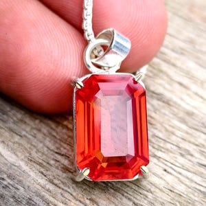 May include: A rectangular, emerald-cut orange-red gemstone pendant set in a silver-toned metal prong setting. The pendant is attached to a silver-toned chain, showcasing a faceted surface that reflects light across its vibrant, translucent body.