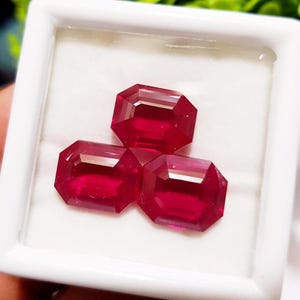 Op de afbeelding: Drie gefacetteerde, rechthoekig geslepen, dieprode edelstenen, gepresenteerd in een witte vierkante doos. De edelstenen hebben een hoge glans en zijn in een driehoek gerangschikt.