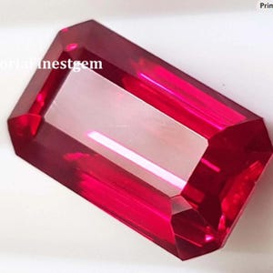 May include: A faceted, rectangular-cut gemstone with a vibrant red hue. The gem has a polished surface that reflects light, creating a brilliant shine. The gemstone is set against a white background. The text "victoriafinestgem" is visible on the gem.