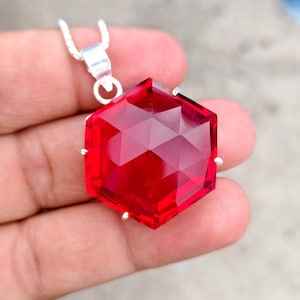 Op de afbeelding: Een zeshoekige hanger met een gefacetteerde, doorschijnende rode edelsteen in een zilverkleurige metalen zetting. De hanger is met een klein, rond oogje bevestigd aan een zilverkleurige ketting en heeft een geometrische slijpvorm die licht reflecteert.