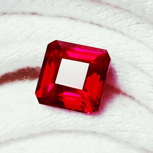May include: A vibrant, square-cut, red gemstone with a white square center. The gemstone is faceted, reflecting light, and set against a soft, white, textured background. The gemstone's color is a deep, rich red.
