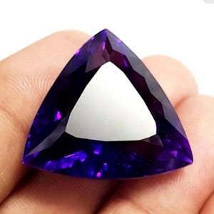 May include: A triangular amethyst gemstone with a deep purple colour and a bright, white centre. The gemstone is held in a hand, highlighting its polished surfaces and the light refraction within the stone.