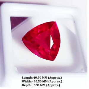 May include: A faceted, triangular ruby gemstone, displayed in a white case. The gem is a vibrant red color. The text below the gem reads: Length: 10.50 MM (Approx.), Width: 10.50 MM (Approx.), Depth: 5.91 MM (Approx.).