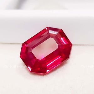 Könnte beinhalten: Ein facettierter Rubin-Edelstein im Radiant-Schliff. Der Edelstein hat eine leuchtend rote Farbe, eine rechteckige Form und abgeschrägte Kanten. Der Edelstein liegt vor einem weißen Hintergrund.