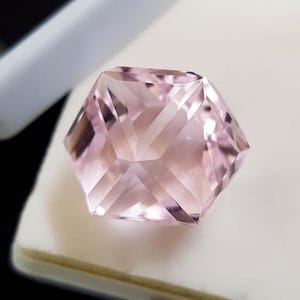 May include: A hexagonal gemstone with a soft pink colour and intricate facets. The cuts create a play of light and shadow, enhancing its brilliance. The gemstone is set against a white background, highlighting its colour and clarity.