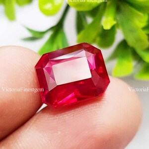 May include: A faceted, rectangular-cut ruby gemstone. The gem is a deep, vibrant red with a lighter, rectangular facet in the center. The gem is held between fingers, with green foliage in the background. The text "VictoriaFinestgem" is visible.
