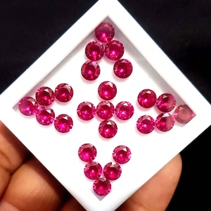 May include: A collection of round, faceted, deep red rubies arranged in a white square tray. The gemstones are displayed against a black background, showcasing their vibrant color and brilliance. The tray is held by a hand.