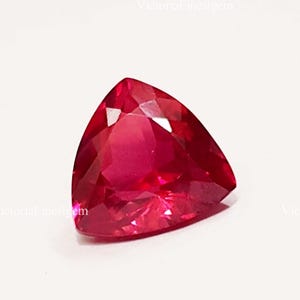 May include: A faceted, triangular-shaped, ruby-red gemstone. The gem has a polished surface with multiple facets that reflect light, creating a brilliant appearance. The gemstone is set against a white background.