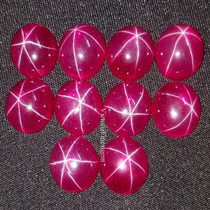 May include: Ten round, polished, star ruby gemstones. Each ruby is a deep pink color with a six-rayed star pattern across the surface. The gemstones are arranged on a dark gray surface.