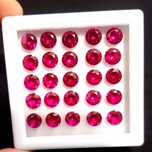 May include: A collection of twenty-one round, faceted, deep red rubies displayed in a white tray. The gemstones are evenly spaced and appear to be of similar size and clarity. The tray is held against a dark background.