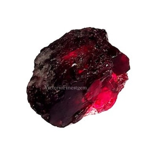 Op de afbeelding: Een ruwe, dieprode granaat edelsteen met een kristallijne structuur. Het ruwe mineraal vertoont een rijke, donkerrode tint met gebieden met hoge glans en lichtbreking. Het oppervlak heeft een textuur.