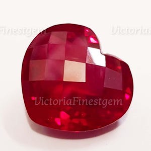 May include: A faceted, heart-shaped gemstone in a deep red hue. The gem has a geometric pattern of cut surfaces, reflecting light. The text "VictoriaFinestgem" is visible on the surface. This gemstone is likely for jewelry making.