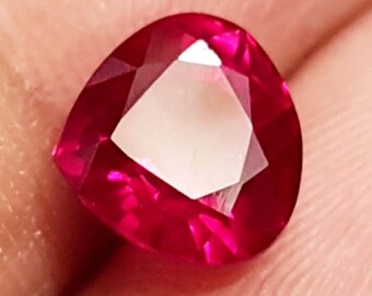 Puede incluir: Una gema de rubí facetada en forma de corazón. La gema es de un rojo intenso y vibrante con una faceta geométrica blanca y brillante en el centro. La piedra se sostiene en una mano, con el tono de la piel que proporciona un cálido contraste con el color de la joya.