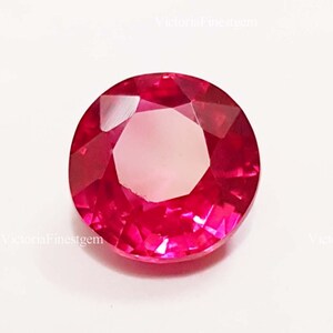 May include: A faceted, round-cut ruby gemstone. The gem is a vibrant shade of red with a bright, reflective surface. The gemstone is set against a white background. The text "VictoriaFinestgem" is visible in the image.