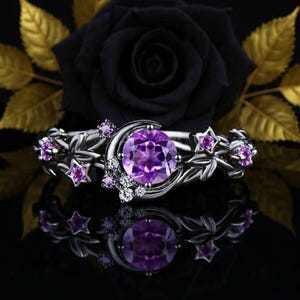 Puede incluir: Un anillo plateado con una gran gema púrpura redonda engastada en un diseño de media luna. El anillo está adornado con detalles en forma de estrella y piedras púrpuras más pequeñas, con un patrón floral y de hojas a lo largo de la banda. El anillo se muestra sobre una superficie negra reflectante.