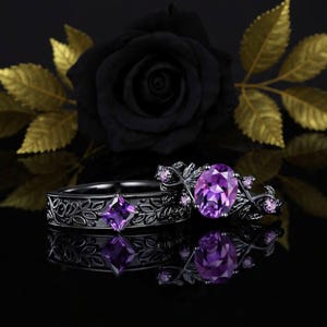 May include: Two black rings with detailed leaf patterns and purple gemstones. One ring has a square gem, the other an oval gem. A black rose and gold leaves are in the background.
