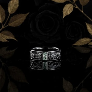 Op de afbeelding: Een donkere, gegraveerde ring met een bladerenpatroon en een rechthoekige, lichtgroene steen. De ring staat tegen een zwarte achtergrond met goudkleurige bladeren en een zwarte roos.