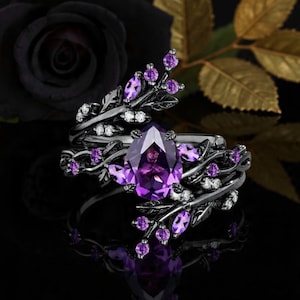 Könnte beinhalten: Ein schwarzer Ring mit einem großen, birnenförmigen lila Edelstein als Mittelpunkt. Der Ring ist mit kleineren lila Steinen und weißen Akzenten verziert, die einem floralen Design ähneln. Der Ring ist vor einem dunklen Hintergrund mit einer schwarzen Rose platziert.