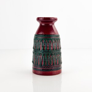 Peut inclure: Un vase en céramique avec une finition rouge profond brillante et un motif texturé vert foncé. Le récipient présente un corps cylindrique qui se rétrécit vers un col évasé, avec des bandes horizontales et des motifs en zigzag verticaux.