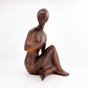 Op de afbeelding: Een bruin, getextureerd beeldhouwwerk van een zittende figuur met een glad, rond hoofd en kort haar. De figuur is afgebeeld in een knielende houding met één hand op de borst en draagt een eenvoudige ketting om de hals.