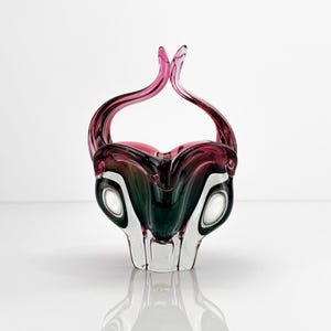Pode incluir: Uma cesta de vidro decorativa com uma base verde-escura que transita para uma borda magenta vibrante. A peça possui duas alças curvas e alongadas que se encontram no topo, criando uma silhueta simétrica com detalhes em vidro transparente.