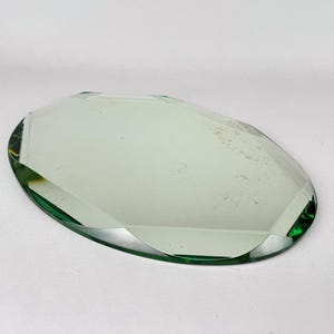 Pode incluir: Um espelho de vidro de formato oval com uma borda chanfrada. A superfície reflexiva tem um tom verde claro, e o perímetro do vidro exibe uma tonalidade verde mais escura onde as facetas chanfradas captam a luz.