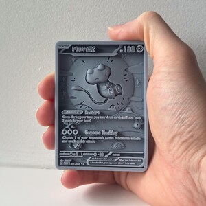 May include: A gray Mew EX Pokémon trading card held in a hand. The card features a 3D embossed design of Mew, with the text "Mew EX" at the top and other details. The card is rectangular and has rounded corners.