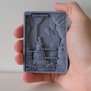 Op de afbeelding: Een grijze 3D-geprinte ruilkaart met een gedetailleerd reliëf van een Charmander-personage in een rotsachtig landschap. De kaart bevat tekst en symbolen en wordt in een hand gehouden.