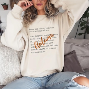 Può includere: Felpa color crema con la parola "Autumn" in un carattere marrone. La felpa presenta un elenco di parole e frasi a tema autunnale, tra cui "cozy jumpers", "pumpkin spice latte" e "harvest skies."