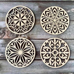 May include: Four laser-cut wooden coasters with intricate floral and geometric designs. Each coaster features a unique pattern, including stylized flowers and symmetrical shapes. The coasters are made of light-coloured wood and are displayed on a wooden surface.