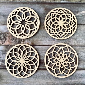 May include: Four circular wooden coasters featuring laser-cut floral and geometric patterns. The coasters are made from light-coloured wood and are displayed on a weathered, grey wooden surface. Each coaster has a unique design.