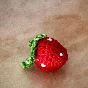 Peut inclure: Une fraise crochetée à la main, de couleur rouge vif, avec de petits détails de graines blanches. La fraise est surmontée d'une tige et de feuilles vertes. Il s'agit probablement d'un objet décoratif ou d'un jouet.