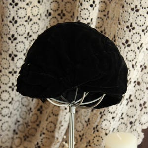 May include: A black velvet hat, possibly vintage, presented on a white metal stand. The hat features a rounded crown and a soft, textured finish. The background includes a lace curtain and a white candle, evoking a classic or antique aesthetic.
