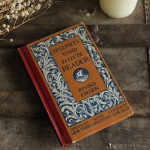 Może przedstawiać: Vintage egzemplarz McGuffey's Third Eclectic Reader, Revised Edition. Książka ma brązową okładkę z niebieskimi i białymi elementami dekoracyjnymi oraz ilustracją statku. Grzbiet jest czerwony, a książka leży na drewnianej powierzchni.