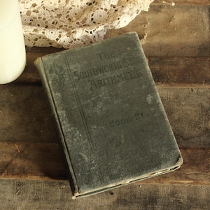 May include: An antique book titled "The Southworth-Stone Arithmetic, Book III" with a faded green cover, resting on a rustic wooden surface. A white candle and lace fabric are in the background, creating a vintage aesthetic.