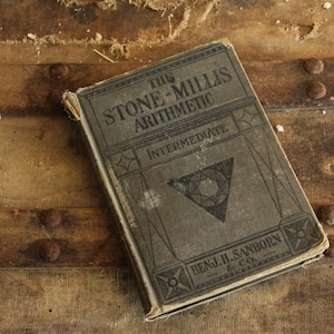 Puede incluir: Un libro antiguo titulado "The Stone-Millis Arithmetic" con "Intermediate" impreso debajo del título. El libro tiene una cubierta gris con diseños geométricos y está sobre una superficie de madera con tachuelas metálicas.