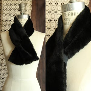 May include: A black faux fur scarf featuring a soft, plush texture and a dark, uniform color. The scarf is secured at the front with a simple, integrated dark elastic band that holds the two ends together, creating a draped, layered appearance.