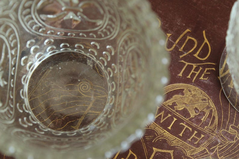May include: Clear glass bowl with ornate, embossed detailing. The bowl's rim has a scalloped edge. The bowl sits atop a brown surface with gold lettering that reads "Und The With".