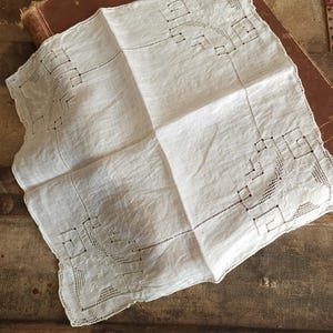 May include: A square white textile napkin featuring intricate openwork embroidery and cutwork details in each corner. The fabric has a visible weave and delicate needlework patterns that form geometric and floral motifs along the edges.