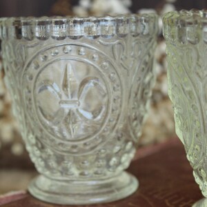 May include: Two clear, vintage-style glass votive candle holders. The holders feature an embossed fleur-de-lis design and ornate detailing. The rims have a scalloped edge. The holders are on a small pedestal base.