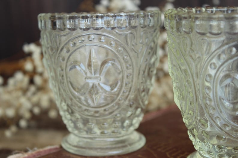 May include: Two clear, vintage-style glass votive candle holders. The holders feature an embossed fleur-de-lis design and ornate detailing. The rims have a scalloped edge. The holders are on a small pedestal base.